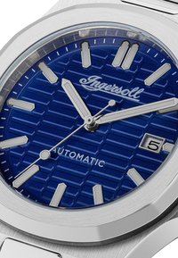 Blauwe hexagoonpatroon horlogekast met zilverkleurige wijzers en markeringen, roestvrijstalen kast en een datumvenster op de 3-uur positie.