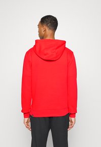 Sweatshirt vermelho com capuz e uma textura suave. Apresenta um bolso canguru e punhos canelados. Costuras visíveis e etiqueta com o logótipo na bainha.