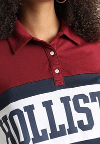 Polo a righe rosse e blu con una banda bianca che presenta la parola "HOLLISTER" in grassetto blu. Collo classico e dettagli con bottoni.