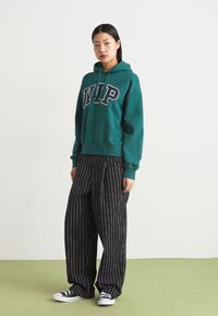 Felpa verde con la scritta "WIP" in lettere nere, abbinata a pantaloni a gamba larga neri a righe e sneaker in tela neri.