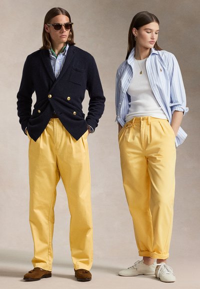 Två modeller bär koordinerade outfits: en i en marinblå dubbelknäppt cardigan och gula byxor; den andra i en randig skjorta och gula byxor.