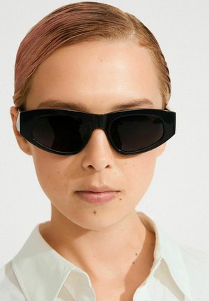 Mustad ülisuured päikeseprillid stiilse nurkjalise raamiga, tumedate läätsede ja valgete külgdetailidega. Kandmisel valge kraega särk.