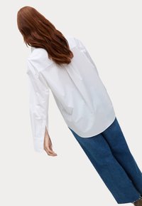Weißes übergroßes Hemd mit strukturiertem Kragen, langen Ärmeln und seitlichen Schlitzen, kombiniert mit blauen Jeans in weitem Schnitt. Glatte Oberfläche.