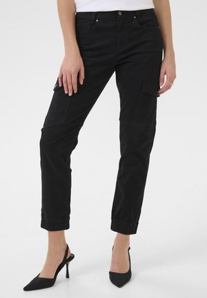 Frau trägt schwarze Cargohosen mit Taschen und schwarze High-Heel Slingback-Schuhe, steht vor einem schlichten Hintergrund.