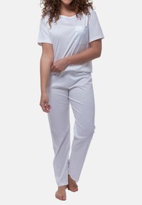 Nainen, jolla on kihara hiukset, pukeutuneena valkoiseen lyhythihaiseen toppiin, jossa on rintatasku, ja matching valkoisiin housuihin, seisoo paljain jaloin yksinkertaisella taustalla.