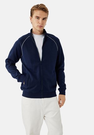 Sudadera navy con cremallera y ribetes blancos, puños y dobladillo de canalé, cuello alto y bolsillos laterales, combinada con pantalones de color claro.