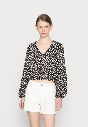 Blusa preta com padrão abstrato em branco e rosa, decote em V e cintura franzida. Combinada com calções de ganga brancos. Mangas longas e bufantes.
