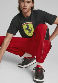 Camiseta negra con el logo amarillo de Ferrari, pantalones de chándal rojos con detalles negros y cremallera, y zapatillas negras con detalles rojos y suelas blancas.