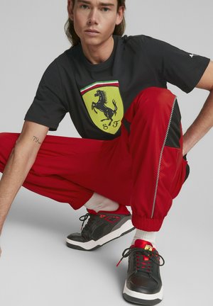 Camiseta negra con el logo amarillo de Ferrari, pantalones de chándal rojos con detalles negros y cremallera, y zapatillas negras con detalles rojos y suelas blancas.