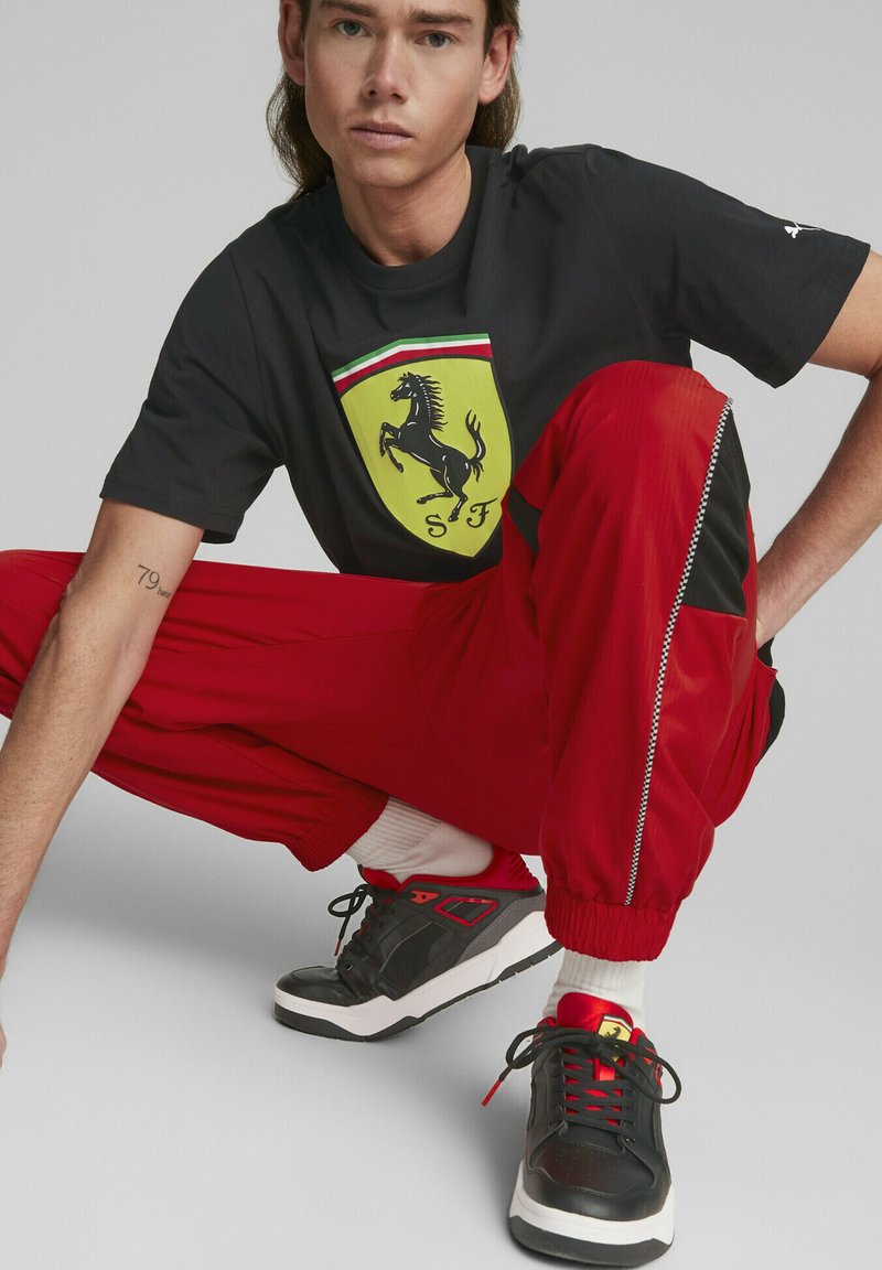 Camiseta negra con el logo amarillo de Ferrari, pantalones de chándal rojos con detalles negros y cremallera, y zapatillas negras con detalles rojos y suelas blancas.