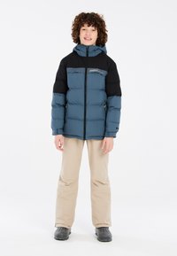 Giacca imbottita blu e nera con cappuccio, dotata di zip frontale e dettaglio del logo. Abbinata a pantaloni beige e stivali grigio scuro.