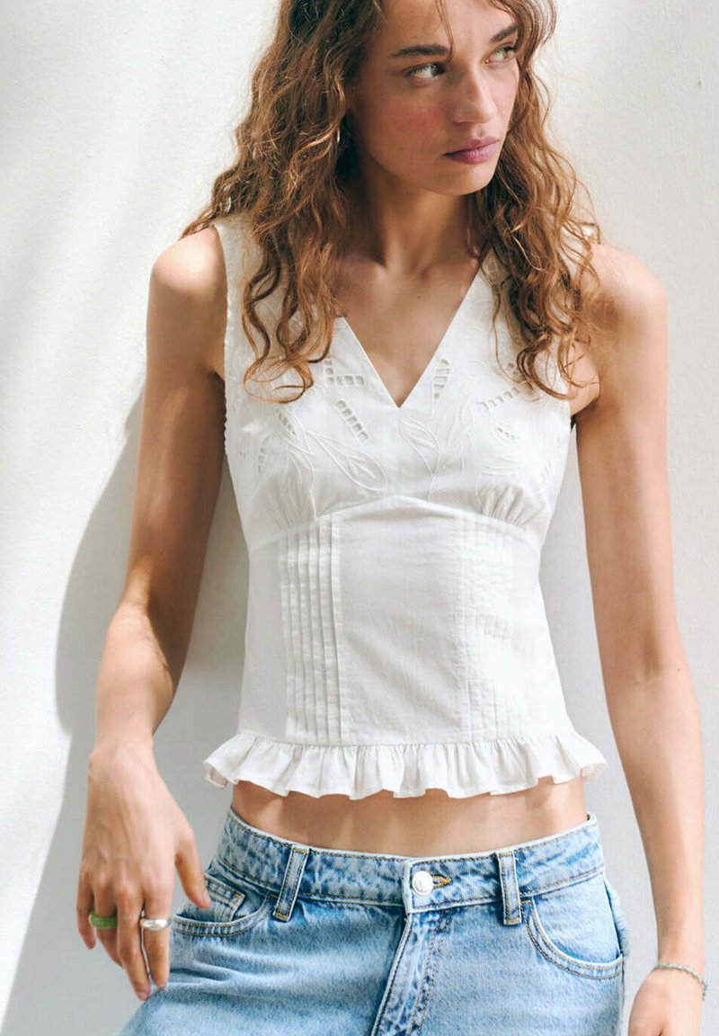 Jeune femme aux cheveux bouclés portant un top blanc brodé sans manches avec un ourlet à volants et un jean taille haute bleu clair contre un mur blanc.