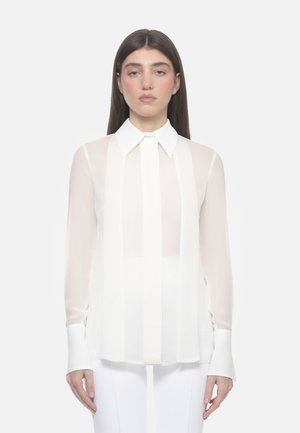 Donna con lunghi capelli castani che indossa una camicetta bianca trasparente con righe verticali e un colletto appuntito, in piedi davanti a uno sfondo semplice.