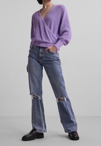 Fioletowy sweter z dzianiny w formie narzutki z głębokim dekoltem w serek i prążkowanymi mankietami, zestawiony z jasnoniebieskimi dżinsami z przetarciami i czarnymi butami na platformie.