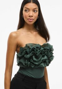 Zelený bezramínkový body s vrstvenými volány a jemným leskem. Hladká, přiléhavá látka s jemným zdrsněním, spárovaná s černými kalhotami.