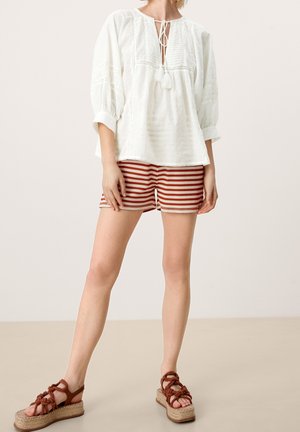 Femme portant une blouse blanche ample avec un lien à pampilles, un short rayé rouge et beige, et des sandales compensées tressées marron, debout sur un sol beige.