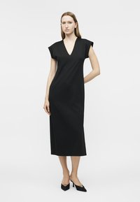 Žena stojaca v čiernych midi šatách bez rukávov s výstrihom do V a čiernych špicatých opätkoch na bielom pozadí.