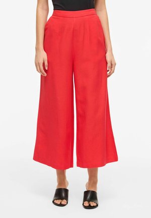 Femme portant un pantalon rouge court taille haute à jambes larges, associé à des sandales noires à bouts ouverts, debout sur un fond blanc.