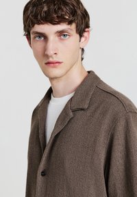 Giovane uomo con capelli castani mossi e occhi blu, indossa una giacca marrone a trama sopra una camicia bianca, mentre guarda direttamente verso la fotocamera.