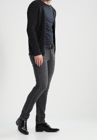 Svart stickad cardigan, marinblå randig skjorta, grå slim fit jeans, blanka svarta läderskor, stående pose, minimal bakgrund.