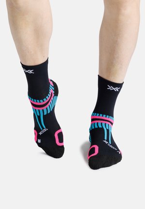 Benen die zwarte sportsokken dragen met blauwe en roze strepen en patronen, staand op een witte achtergrond.