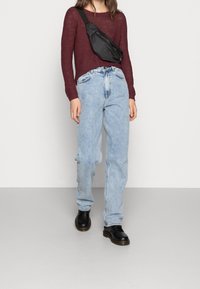 Pull en maille bordeaux, jean baggy bleu clair et bottes noires. Un sac bandoulière noir est porté en travers du torse.
