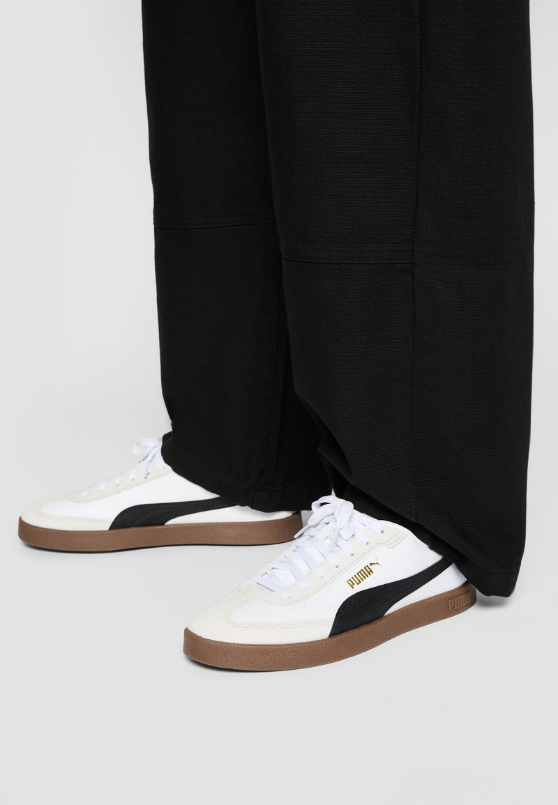 Witte en zwarte sneakers met een gestructureerd ontwerp, voorzien van een bruine rubberen zool en gouden merkvoering aan de zijkant. Zwarte broek bedekt een deel van de schoenen.