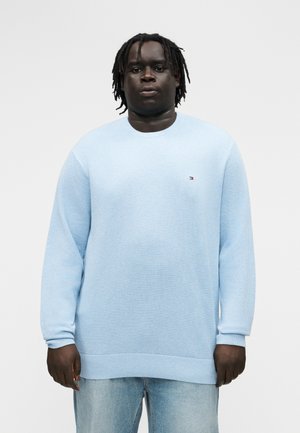 Νέος άνδρας με ντραντλόκς, φοράει ανοιχτό γαλάζιο πουλόβερ με μακριά μανίκια και ανοιχτό γαλάζιο τζιν, κοιτάει προς τα εμπρός με φόντο ένα απλό υπόβαθρο.