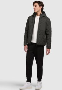Black quilted jacket with hood, featuring a zippered chest pocket and subtle geometric texture. Worn over a white shirt with black pants.
