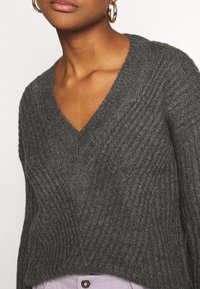 Maglione grigio lavorato a maglia con scollatura a V e texture a costine. Presenta una vestibilità comoda e cuciture visibili, abbinato a pantaloni di colore chiaro.