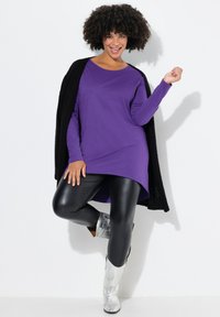Lila Langarmoberteil mit abgerundetem Saum, kombiniert mit glänzenden schwarzen Leggings und silbermusterierten Ankle Boots, ergänzt durch einen schwarzen Cardigan.