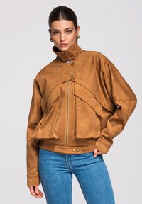 Femme aux cheveux lissés en arrière, portant une veste en suède beige à col montant avec de grandes poches à rabat et un jean bleu, posant devant un fond uni.