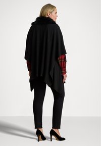 Poncho negru cu guler din blană artificială, design stratificat și tiv asimetric; purtat peste o cămașă cu carouri roșii și asortat cu blugi negri skinny și tocuri.