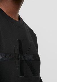 Musta collegepaita pyöreällä kaula-aukolla, jossa on brodeerattu logo ja teksturoitua nauhakoristelua rinnan kohdalla. Sujuva kankaan pinta.