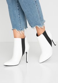 Botines blancos con un diseño elegante, punteras afiladas, altos tacones de aguja y paneles laterales elásticos negros. Terminados con una textura suave.