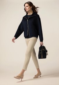 Marineblauwe cropped jas met gouden ritsen, gecombineerd met een donkere trui, beige slim-fit broek en beige hakken. Houdt een zwarte handtas met gouden accenten vast.