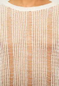 Närbild av en persons övre rygg som bär en vit, genomskinlig, randig stickad topp med vertikala mönster av öppen väv.