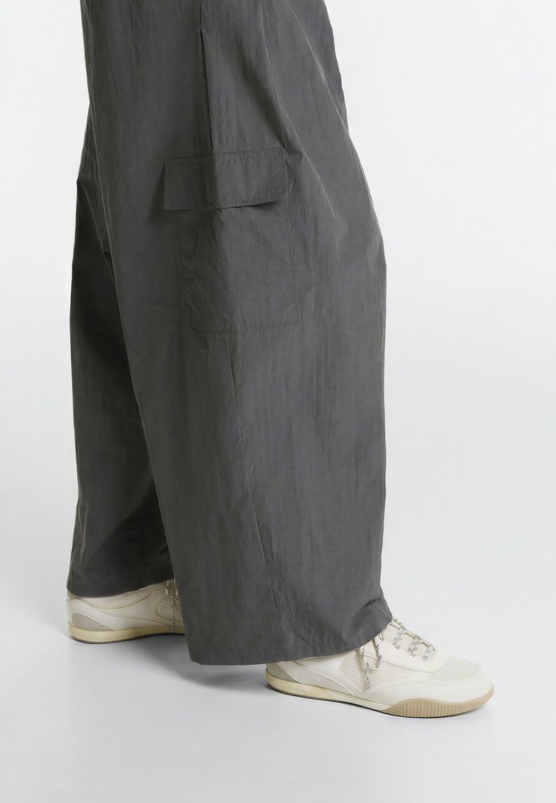 Personne portant un pantalon ample gris foncé et des baskets blanches, debout sur un sol gris clair avec un fond uni.