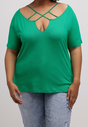 Groen, kortermig shirt met een diepe V-hals en gekruiste bandjes bij de schouders. Zachte stof; gecombineerd met lichtblauwe, versleten jeans.