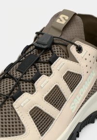 Gros plan sur une chaussure de trail Salomon marron et beige avec des panneaux en maille et un système de laçage noir à dégagement rapide sur fond blanc.