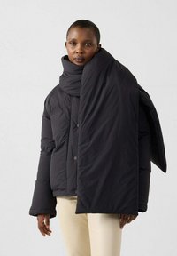 Zwarte gewatteerde jas met een grote, vallende kraag en oversized mouwen. Voorzien van een knoopsluiting en een gladde, matte textuur.