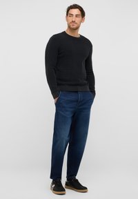 Schwarzer gerippter Strickpullover, blaue lässige Jeans und schwarze Sneaker mit hellbrauner Sohle. Das Model steht vor einem schlichten Hintergrund.