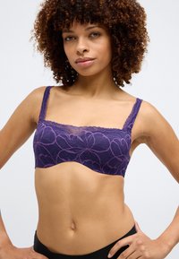 Violeta mežģīņu krūšturis ar ziedu rakstu, viļņotām malām un platām siksnām. Dizainam ir gluda tekstūra un bezšuvju siluets.