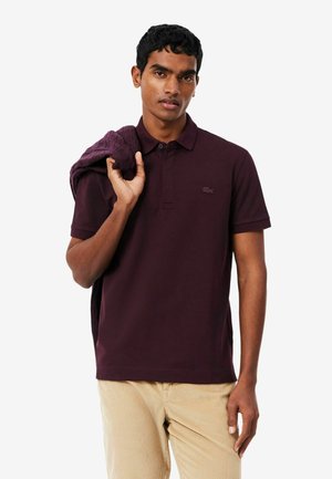 Hombre que lleva una camisa polo de color púrpura oscuro y pantalones beige sostiene un suéter oscuro doblado sobre su hombro contra un fondo blanco liso.