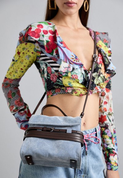 Femeie îmbrăcată cu un top scurt din petice colorate cu flori, blugi de culoare albastră deschisă și o geantă crossbody din denim deschis cu bretele de culoare maro închis.
