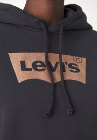 Gros plan sur un sweat à capuche noir avec un patch rectangulaire en bronze brillant affichant le logo Levi's et des cordons noirs.
