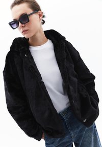 Zwarte faux fur jas met een capuchon, voorzien van een zachte textuur en knoopsluitingen, gedragen over een wit T-shirt en blauwe high-waisted jeans.