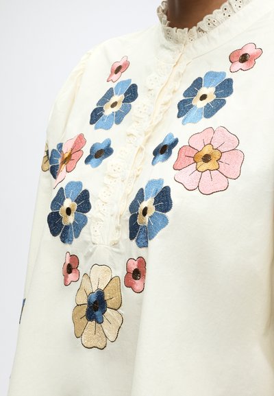 Blouse crème avec de grandes et petites fleurs brodées en bleu, rose et beige autour de la poitrine et des épaules, avec col et patte de boutonnage ornés de dentelle.