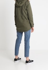 Olivgrön parkas med huva och fuskpäls, kombinerad med ljusblå raka jeans och svarta sneakers. Bakifrån, inga synliga detaljer.