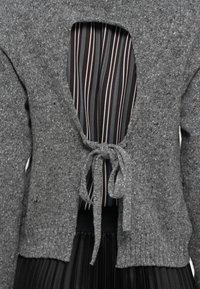 Cardigan grigio a maglia annodato davanti su una blusa a righe e una gonna pieghettata nera, che mostra i dettagli di un outfit a strati.